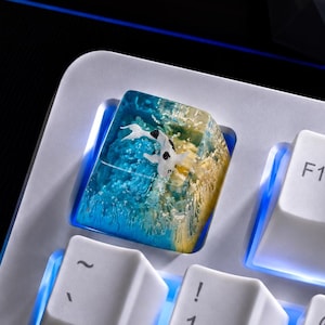 May include: A custom resin mechanical keyboard keycap featuring a miniature koi fish swimming in a gradient of blue and yellow water. The keycap is installed on a white keyboard with blue backlighting, positioned next to the F1 key and number keys.