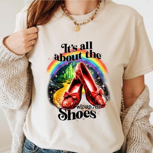 May include: A cream-coloured t-shirt with a Wizard of Oz graphic. The design shows a rainbow, the Emerald City, and red sequined shoes with bows. The text reads "It's all about the Shoes."