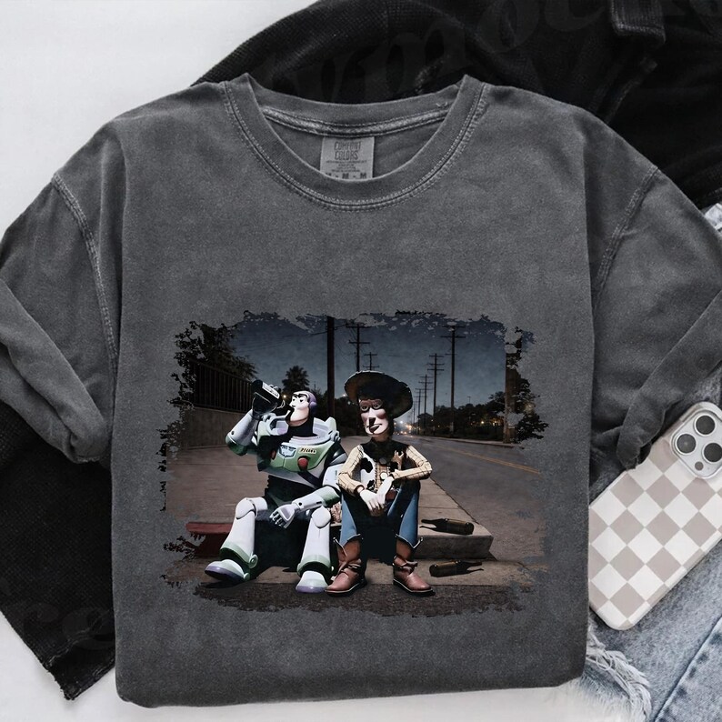 A gray t-shirt with a printed image of two characters from the Toy Story franchise, Buzz Lightyear and Woody, sitting on a bench and holding a beer bottle.