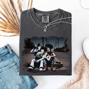 a gray t-shirt with a printed scene of two characters from the Toy Story franchise, including Buzz Lightyear and Woody, sitting on a bench and holding drinks.