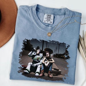 a blue t-shirt with a printed image of two characters from the Toy Story franchise, Buzz Lightyear and Woody, sitting on a bench and holding a camera.