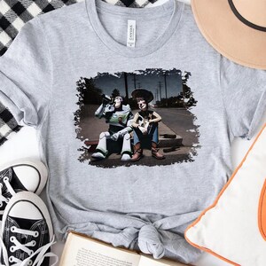a gray t-shirt with a printed image of two characters from the Toy Story franchise, Buzz Lightyear and Woody, sitting on a curb.