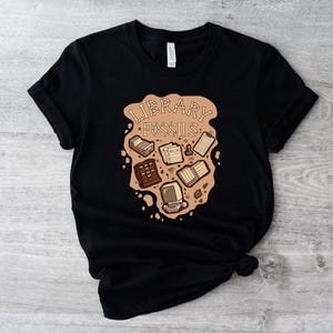 May include: Black t-shirt featuring a brown graphic with the words "LIBRARY FOSSILS" and illustrations of books, a computer, and other library-related items. The design has a vintage, fossil-like aesthetic.