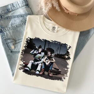 a white t-shirt with a printed graphic of two characters from the Toy Story movie, Buzz Lightyear and Woody, sitting on a bench. The t-shirt is placed on a white surface, along with a beige hat and a pair of blue jeans.