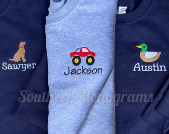 Puede incluir: Tres sudaderas de manga larga con cuello redondo. Dos son azul marino y una es gris jaspeado. Cada una presenta un gráfico bordado: un perro marrón sobre el nombre Sawyer, un camión monstruo rojo sobre Jackson y un pato real sobre Austin.