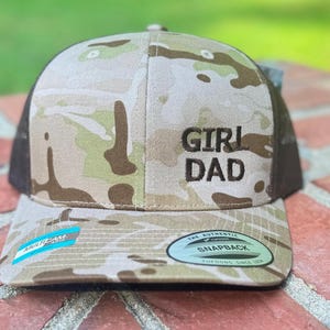 May include: A camouflage print baseball cap with the text "GIRL DAD" in black. The cap features a brown mesh back and a curved brim. The hat has a snapback closure and a blue and white tag that says "MULTICAM".