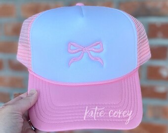 Puede incluir: Una gorra de camionero rosa y blanca con un lazo rosa bordado en la parte delantera. La gorra tiene una malla rosa en la parte posterior y las palabras "katie corey" están bordadas en la visera rosa.