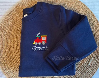 Puede incluir: Sudadera azul marino con un tren bordado en rojo, amarillo y verde y el nombre "Grant" debajo.