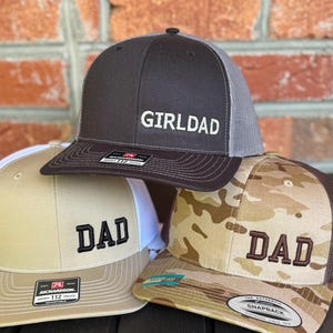 May include: Three baseball caps with the word "DAD" or "GIRLDAD" embroidered on the front. One cap is brown with a gray mesh back, another is tan with a white mesh back, and the third is camouflage with a brown mesh back.