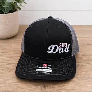 May include: A black trucker hat with grey mesh back panels and an adjustable snapback closure. The front features embroidered text that reads Girl Dad in white and light pink script. A black and white Richardson 112 sticker is on the brim.