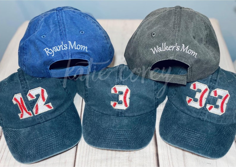 May include: Six baseball caps, three blue, two black, and one gray. The blue caps have white stitching and red numbers, 12, 3, and 33. The gray cap has white stitching and the text "Walker's Mom". The blue cap in the back has white stitching and the text "Ryan's Mom".
