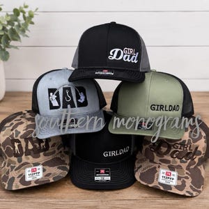 May include: A collection of trucker hats in various colors and patterns. The hats feature the text "GIRL DAD" or "GIRLDAD" in white script. Some hats have fishing-themed graphics. The hats have a mesh back and a curved brim.