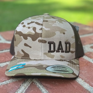 May include: Camouflage baseball cap with the word "DAD" embroidered in brown. The cap features a mesh back and a curved brim. The front of the cap has a camouflage pattern in shades of tan, brown, and green.
