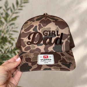 May include: A camouflage baseball cap with the words "Girl Dad" embroidered in brown. The hat has a brown mesh back and a curved brim. A white tag with black text is on the front.