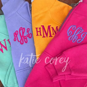 May include: Four quarter-zip sweatshirts in mint green, lavender, yellow, and hot pink. Each sweatshirt features a unique embroidered monogram on the chest. The mint green has pink letters, the lavender has pink script, the yellow has pink block letters, and the hot pink has lavender script.