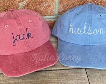 May include: Two personalized baseball caps, one red with the name "jack" embroidered in navy blue, and one blue with the name "hudson" embroidered in white.