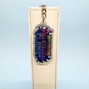 May include: A holographic keychain featuring the text "FLAMES PHANTASMAL" and "MEGA EVOLUTION" within a flame-shaped design. The keychain is suspended from a silver chain and set against a light backdrop.