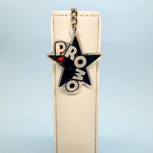 May include: A star-shaped keyring with the word "PROMO" in white letters. The star has a holographic, iridescent finish and is attached to a silver chain. The background is a light blue colour.