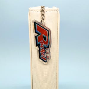 May include: A clear acrylic keyring featuring the red and blue text Team Rocket in a stylised font, attached to a silver metal chain. The keyring is hanging against the side of a vertical, textured, off-white rectangular object.