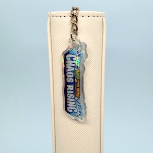 May include: A clear, iridescent keychain with the text "Chaos Rising" in blue and white, with "Mega Evolution" in orange. The keychain is attached to a silver chain.