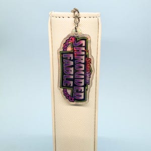 Könnte beinhalten: Ein klarer Acryl-Schlüsselanhänger mit dem Text "SHROUDED FABLE" und "Scarlet & Violet" in einem farbenfrohen Design. Der Schlüsselanhänger ist an einer silbernen Kette befestigt und hängt vor einem weißen Objekt.