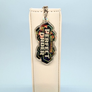 May include: A holographic keychain with the words "PERFECT ORDER" in a stylized font. The keychain has a silver chain and a geometric design with the text "MEGA EVOLUTION".