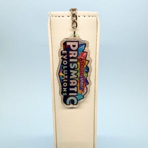 Puede incluir: Un llavero acrílico transparente con una cadena plateada. El llavero presenta el texto "Prismatic Evolutions" y "Scarlet & Violet" en un diseño geométrico colorido. El llavero cuelga sobre un fondo blanco.