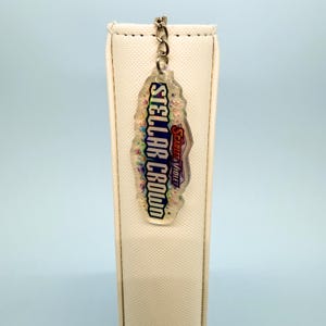 May include: A clear, irregular-shaped keychain with the text "STELLAR CROWD" in a colorful font. The keychain also features the text "SCARLET & VIOLET" in a smaller font. A silver-colored chain connects the keychain to a silver-colored ring.