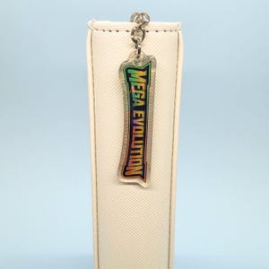 May include: A clear, rectangular keychain with the text "MEGA EVOLUTION" in green, yellow, and orange. The keychain is attached to a silver chain and hangs in front of a white, textured surface.