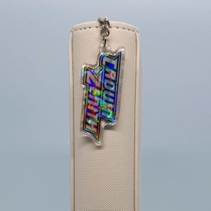 May include: A clear acrylic keychain with the text "Trouth Day" in a holographic, multi-colored design. The keychain is attached to a silver-colored metal chain and is hanging in front of a beige, textured background.