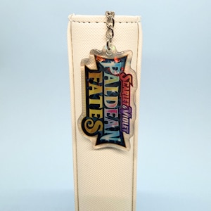 May include: A clear acrylic keychain with the text "Paldean Fates" and "Scarlet & Violet" in a colorful design. The keychain is attached to a silver chain and is hanging in front of a white background.