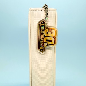 May include: A cream-coloured textured binder featuring a hanging acrylic keychain. The keychain is shaped like a badge with a gold and black border, displaying the text 30th CELEBRATION in bold yellow and orange lettering with a small red circular icon.