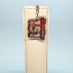 May include: A clear acrylic keyring featuring the number "151" in red and silver, and the words "Scarlet & Violet" in purple. The keyring is suspended from a silver chain against a cream-coloured backdrop.