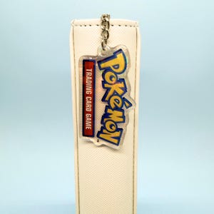 May include: A cream-colored binder with a textured fabric cover featuring a hanging acrylic keychain. The keychain displays the yellow and blue Pokémon Trading Card Game logo with a holographic finish, attached by a silver metal chain.