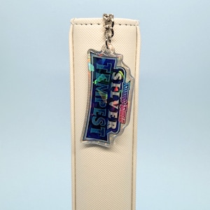 May include: A clear, iridescent keychain with the text "SILVER TEMPEST" in blue and white, and the "SWORD & SHIELD" logo. The keychain is attached to a silver chain and hangs in front of a white, cylindrical object.