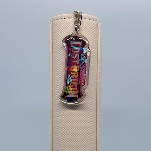 Könnte beinhalten: Ein klarer Acryl-Schlüsselanhänger mit dem Text "Lost Origin" und "Swords & Shield" in einem farbenfrohen Design. Der Schlüsselanhänger ist an einer silbernen Kette befestigt und hängt vor einem beigen, strukturierten Hintergrund.
