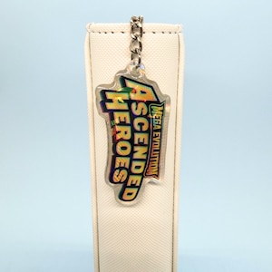 May include: A clear acrylic keychain with the words "ASCENDED HEROES" in green and yellow, and "MEGA EVOLUTION" in white. The keychain is attached to a silver chain. The background is white.