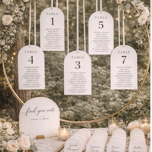 Puede incluir: Plano de mesa de boda con los números de mesa 1, 3, 4, 5 y 7. Cada tarjeta de mesa blanca tiene una parte superior redondeada y nombres de invitados. Un letrero dice "find your seat" con los nombres Emma & James.
