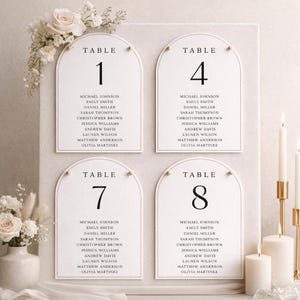 Puede incluir: Cuatro tarjetas de números de mesa de boda con la parte superior arqueada, cada una mostrando un número de mesa y nombres de invitados en fuente negra. Las tarjetas son blancas con un borde dorado y están decoradas con arreglos florales y velas.