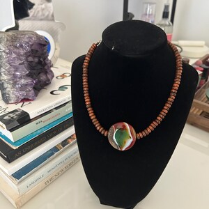 Puede incluir: Un collar artesanal con un colgante central redondo con diseños verdes, rojos y blancos. El colgante está ensartado en una serie de pequeñas cuentas de madera de color marrón rojizo. El collar se exhibe sobre un busto de terciopelo negro.