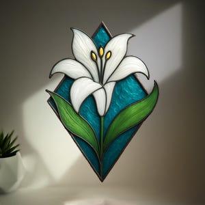 May include: Stained glass art piece featuring a white lily flower with yellow accents, set against a turquoise diamond-shaped background. The lily has green leaves and a dark brown border. A decorative floral design for home decor.