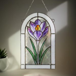May include: Stained glass window panel featuring a purple crocus flower with green leaves. The flower has a yellow center and is set against a textured glass background. The panel is framed in white and hangs from a silver chain.