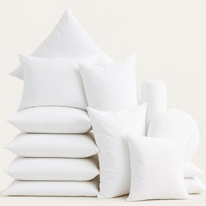 May include: An assortment of white pillows in various shapes, including square, rectangular, triangular, cylindrical, and round. The pillows are arranged together, creating a soft and inviting display. The background is plain.