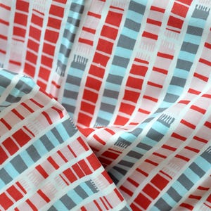 May include: Close-up of a fabric featuring a repeating pattern of red, grey, and light blue rectangles on a white background. The fabric is folded, revealing the pattern's continuity and texture. The design has a modern, geometric style.