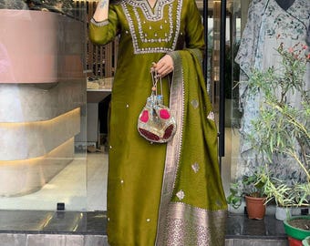 May include: Olive green embroidered salwar kameez suit with matching dupatta. The suit features intricate embroidery around the neckline and cuffs. A small, decorative purse with red and gold accents is held in the hand.
