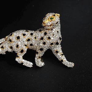 Puede incluir: Broche de leopardo dorado, incrustado con cristales transparentes y manchas de esmalte negro. El leopardo tiene ojos verdes y está representado en posición de caminar. El broche está sobre un fondo negro.