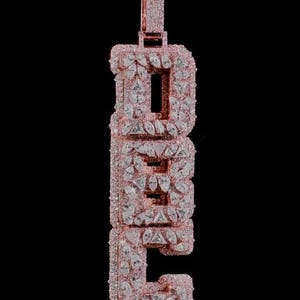 Puede incluir: Un colgante de oro rosa con forma de letras D, E, C apiladas verticalmente. La superficie de las letras está cubierta por numerosas piedras transparentes de talla pera y triangular, con una anilla rectangular en la parte superior.