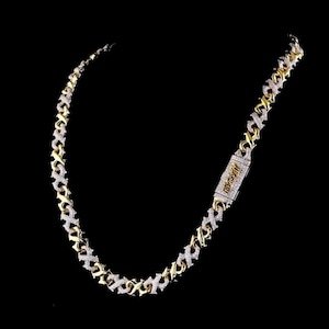 May include: A gold-coloured chain necklace featuring an X-shaped pattern, with white, sparkling stones. The necklace has a rectangular clasp with text. The background is black.
