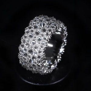 May include: A dazzling, silver ring completely encrusted with numerous, clear gemstones. The ring's surface is covered in closely set, sparkling stones, creating a brilliant, textured appearance. The ring is set against a dark background, highlighting its reflective qualities.