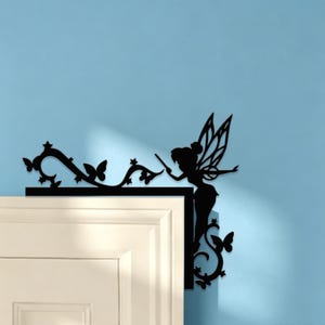 May include: A black silhouette wall decal designed for a door corner, featuring a fairy with wings holding a wand, surrounded by swirling vine patterns and butterfly shapes. The decorative piece is positioned on the upper right corner of a white door frame.